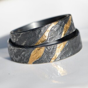 May include: Two black and gold wedding bands with a textured finish. The bands are stacked on top of each other and have a gold stripe design.