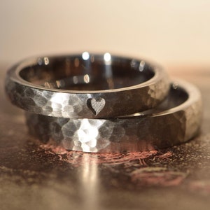 May include: Two silver hammered wedding bands, one with a small heart cutout. The bands are stacked on top of each other.
