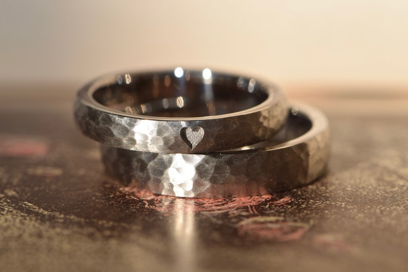 May include: Two silver hammered wedding bands, one with a small heart cutout. The bands are stacked on top of each other.