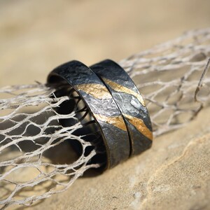 May include: Two black wedding bands with gold accents. The bands are textured and have a hammered finish. The bands are lying on a piece of driftwood.