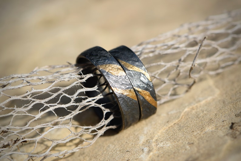 May include: Two black wedding bands with gold accents. The bands are textured and have a hammered finish. The bands are lying on a piece of driftwood.