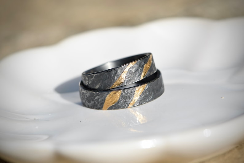 May include: Two black and gold wedding bands with a textured finish. The bands are stacked on top of each other and have a gold stripe design.