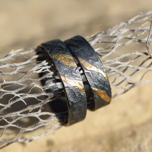 May include: Two black wedding bands with gold accents. The bands have a textured surface and are stacked on top of each other.