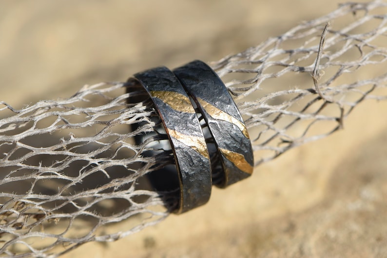 May include: Two black wedding bands with gold accents. The bands have a textured surface and are stacked on top of each other.