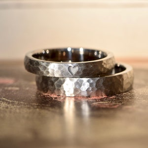 May include: Two silver hammered wedding bands, one with a small heart engraved on the inside. The bands are stacked on top of each other.