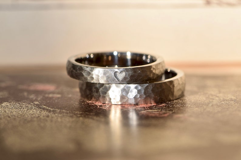 May include: Two silver hammered wedding bands, one with a small heart engraved on the inside. The bands are stacked on top of each other.