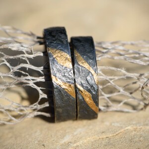 May include: Two black wedding bands with gold accents. The bands have a textured surface and are stacked on top of each other.
