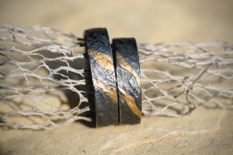 May include: Two black wedding bands with gold accents. The bands have a textured surface and are stacked on top of each other.