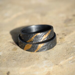 May include: Two black wedding bands with gold accents. The bands have a textured surface and are stacked on top of each other.