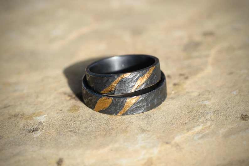 May include: Two black wedding bands with gold accents. The bands have a textured surface and are stacked on top of each other.