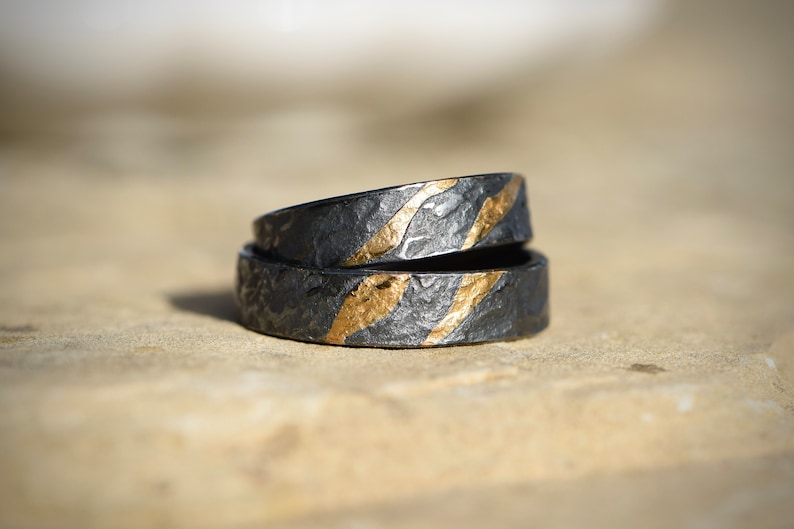 Eheringe schwarz mit Elementen aus Roségold und mit gehämmerter Oberflächenstruktur, welche die Strömung eines Flusses symbolisieren soll.Diezwei Goldelemente stehen für zwei Leben im selben Fluss , jedoch steht jeder stabil für sich.