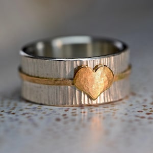 May include: A silver band with a textured finish and a gold heart-shaped accent. The band has a thin gold stripe around the center.