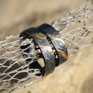May include: Two black and gold wedding bands with a hammered texture. The bands are stacked on top of each other and have a thin gold stripe running around the edge.