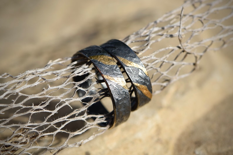 May include: Two black and gold wedding bands with a hammered texture. The bands are stacked on top of each other and have a thin gold stripe running around the edge.