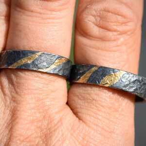 May include: Two silver rings with a textured finish and thin gold stripes. The rings are worn on a person's finger.