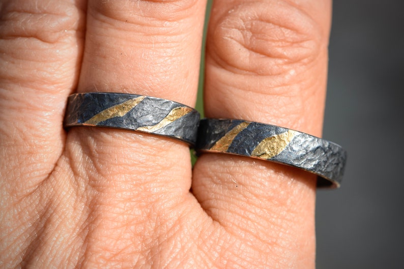 May include: Two silver rings with a textured finish and thin gold stripes. The rings are worn on a person's finger.