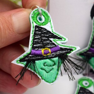 May include: Green witch face with black hat and purple ribbon, embroidered on white felt with black fringe.