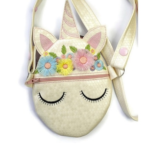 May include: A white unicorn-shaped crossbody bag with a pink zipper, embroidered flowers, and a pink and white striped horn. The bag has a white strap with a pink button.