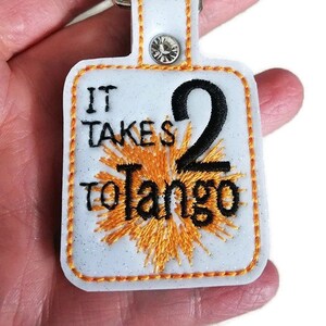 以下が含まれることがあります： オレンジのグリッターと黒い刺繍が施された白いキーホルダーで、「タンゴには2人必要」と書かれています。