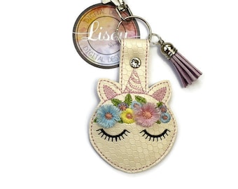 May include: A white unicorn keyring with a floral crown, pink tassel, and a silver charm. The keyring is attached to a silver key ring with a silver clasp.