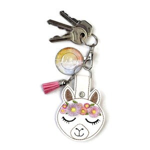 May include: White llama keychain with a pink tassel, a flower crown, and closed eyes. The keychain has a silver ring and a silver clasp. The keychain is attached to a set of keys.