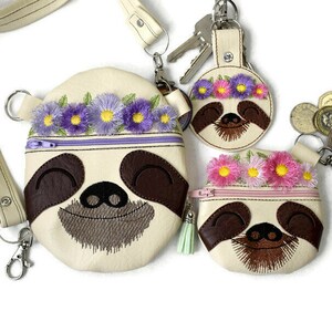 Könnte beinhalten: Drei slothförmige Reißverschlusstaschen mit gestickten Blumenkränzen. Die größte Tasche hat einen Schultergurt und einen silbernen Verschluss. Die kleinste Tasche ist an einem silbernen Schlüsselring befestigt.