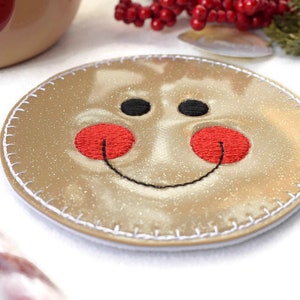 May include: A round, gold-colored coaster with a stitched edge. The coaster has a smiling gingerbread man face embroidered on it with black eyes, a black smile, and red cheeks.
