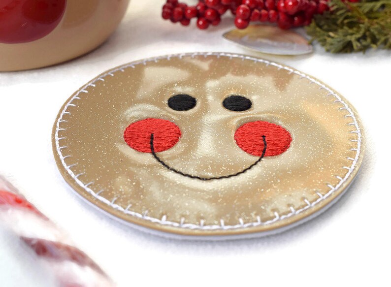May include: A round, gold-colored coaster with a stitched edge. The coaster has a smiling gingerbread man face embroidered on it with black eyes, a black smile, and red cheeks.
