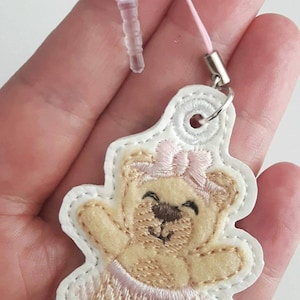 May include: A white phone charm with a brown teddy bear wearing a pink bow and a pink tutu. The charm is attached to a pink string and a clear plastic phone holder.