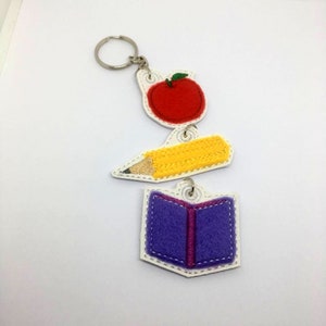 May include: A red apple, yellow pencil, and purple book charm keyring. The charms are attached to a silver key ring.