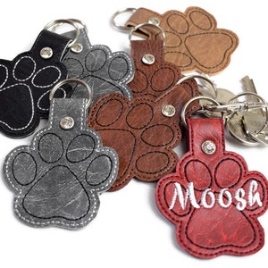 May include: Six leather paw print keyrings in various colours, including black, grey, brown, and red. One keyring has the name "Moosh" embroidered on it.