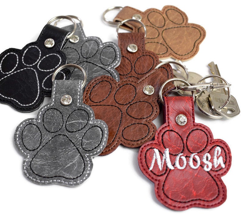 May include: Six leather paw print keyrings in various colours, including black, grey, brown, and red. One keyring has the name "Moosh" embroidered on it.