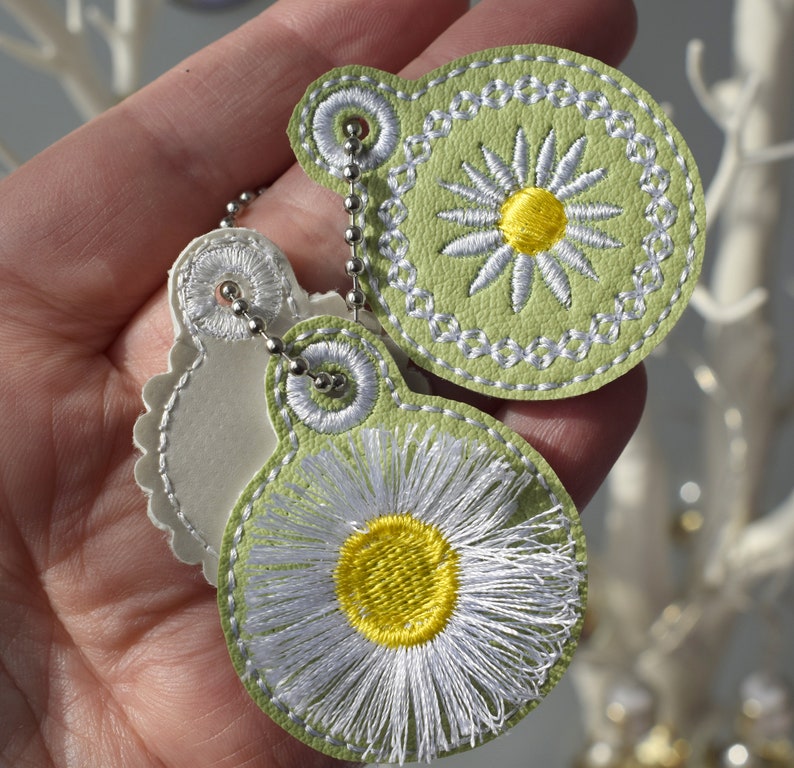 May include: Two green leather keyrings with white daisy embroidery. The keyrings are attached to a silver chain.