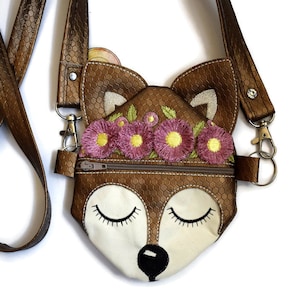 May include: A small brown faux leather crossbody bag with a zip closure. The bag is shaped like a fox's face with embroidered pink flowers and green leaves around the head. The fox has closed eyes and a black nose.
