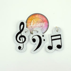 May include: Three white fabric keychains with black embroidered musical notes. The keychains feature a treble clef, a bass clef, and a quarter note.