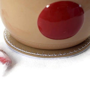 May include: A brown ceramic mug with a red circle design sits on a brown and white coaster. The mug has a handle and is partially visible.