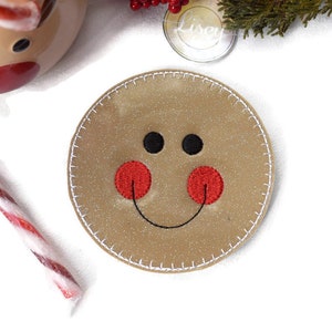 May include: A brown gingerbread man-shaped coaster with a white stitched border. The gingerbread man has black eyes, a red nose, and a stitched smile.