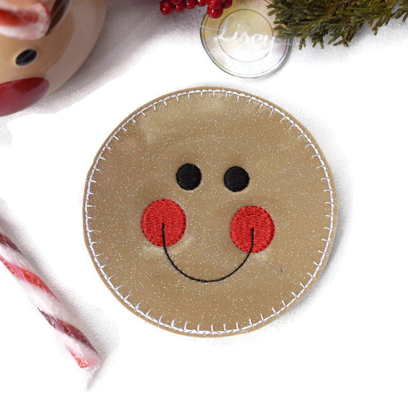 May include: A brown gingerbread man-shaped coaster with a white stitched border. The gingerbread man has black eyes, a red nose, and a stitched smile.