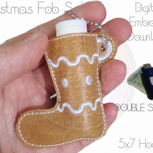 May include: A brown gingerbread boot-shaped fob with white embroidery, designed to hold a small lip balm or other small item. The fob has a silver chain attached to it. The image also shows a USB drive and a lip balm tube, suggesting the fob is a digital embroidery download.