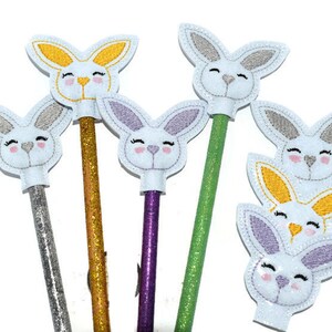 May include: Six pencil toppers featuring white felt bunny faces with embroidered details. The bunnies have different colored ears, including purple, yellow, and gray. The pencils are covered in glitter and come in silver, gold, green, and purple.