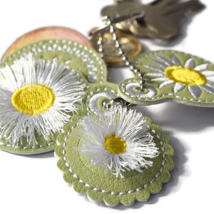 May include: A green leather keychain with three embroidered white daisies with yellow centres. The keychain is attached to a silver chain.