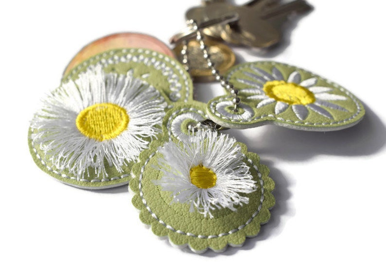 May include: A green leather keychain with three embroidered white daisies with yellow centres. The keychain is attached to a silver chain.