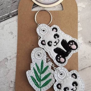 May include: A white keychain with three charms: a green leaf, a panda bear, and another panda bear. The keychain is attached to a silver ring. The charms are embroidered with white thread and have black and pink details. The background is a brown paper tag with the text "Lisey Designs One of a Kind" printed on it.