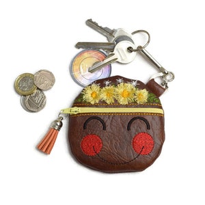 May include: A brown leather coin purse with a zip closure and a tassel. The purse is decorated with a smiling face and a flower crown. The purse is attached to a silver keyring with keys.