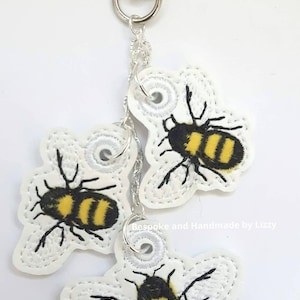 May include: A white keychain with three embroidered bumblebees. The bees are yellow and black with white wings. The keychain has a silver chain and a silver clasp.