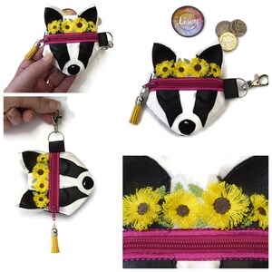 Pu&ograve; includere: Un portamonete a forma di tasso bianco, nero e rosa, con un nappa gialla e una cerniera. Il tasso ha una corona di girasoli e gli occhi chiusi. Ha un cinturino nero e una chiusura argentata.