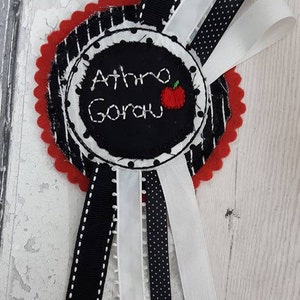 May include: A red and white fabric circle with black stitching and the text "Athro Gorau" with a red apple embroidered on it.  The circle is attached to five black and white ribbons.