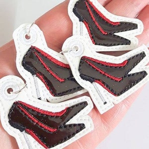 May include: Three white fabric keychains with black and red embroidered high heel shoes. The keychains are designed to look like a pair of shoes.