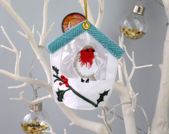 May include: A white felt Christmas ornament shaped like a birdhouse with a robin inside. The birdhouse has a blue and white striped roof and a clear plastic window. There are red berries and green holly leaves on a branch in front of the robin.