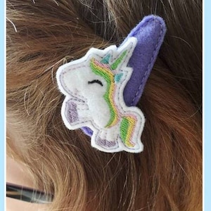 May include: A purple felt hair clip with a white unicorn embroidered on it. The unicorn has a rainbow mane and tail.